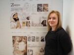 Uczennica stoi przy swojej pracy dyplomowej z projektowania graficznego zawieszonej na ścianie auli szkolnej. Kliknięcie w miniaturkę obrazka spowoduje wyświetlenie powiększonego zdjęcia.