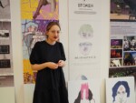 Uczennica stoi przy swojej pracy dyplomowej z projektowania graficznego zawieszonej na ścianie auli szkolnej. Kliknięcie w miniaturkę obrazka spowoduje wyświetlenie powiększonego zdjęcia.