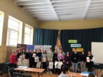 Uczniowie i nauczyciele stoją i pozują do zdjęcia na tle zielonej kotary w sali gimnastycznej. Uczestnicy konkursu trzymają dyplomy i papierowe torby z nagrodami. Kliknięcie w miniaturkę obrazka spowoduje wyświetlenie powiększonego zdjęcia.